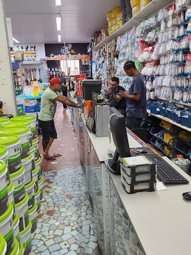 Funcionários atendendo um cliente no balcão, com latas de tinta e parede de ferragens ao redor
